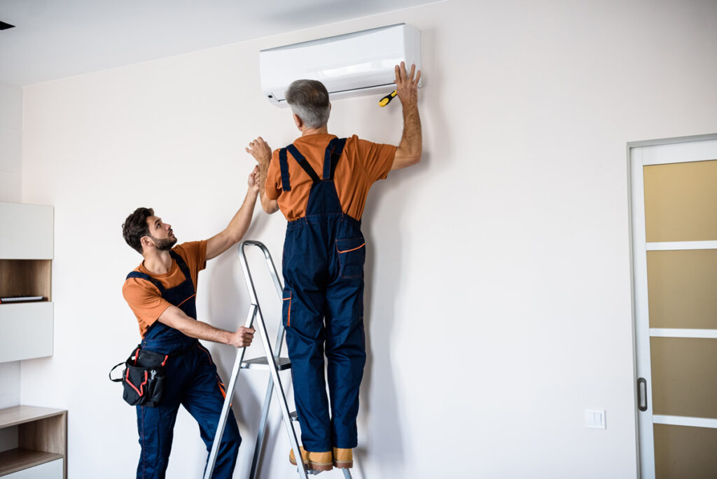 Two professionals in orange shirts installing an AC unit on the wall of a North Fort Myers home.