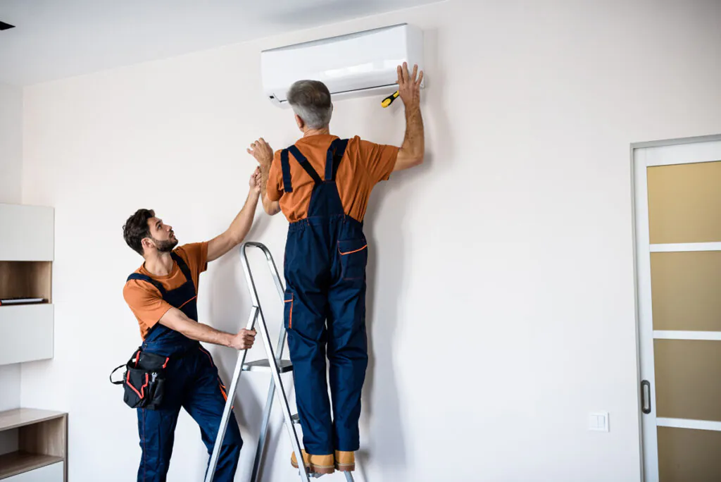 Two professionals in orange shirts installing an AC unit on the wall of a North Fort Myers home.